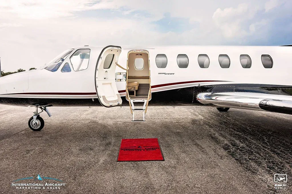 The open main entry door and airstairs of a white Cessna Citation business jet with a red welcome mat on the tarmac.