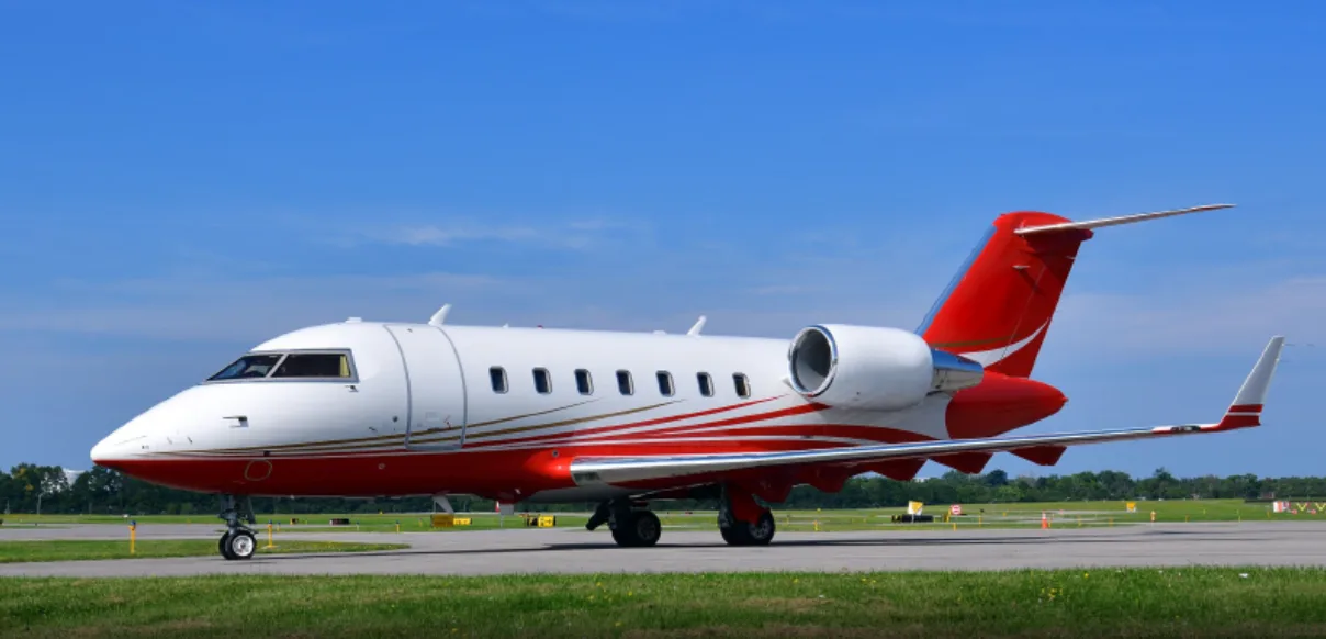 A white business jet with red and gold stripes on the fuselage and tail, parked on an airfield under a blue sky.
