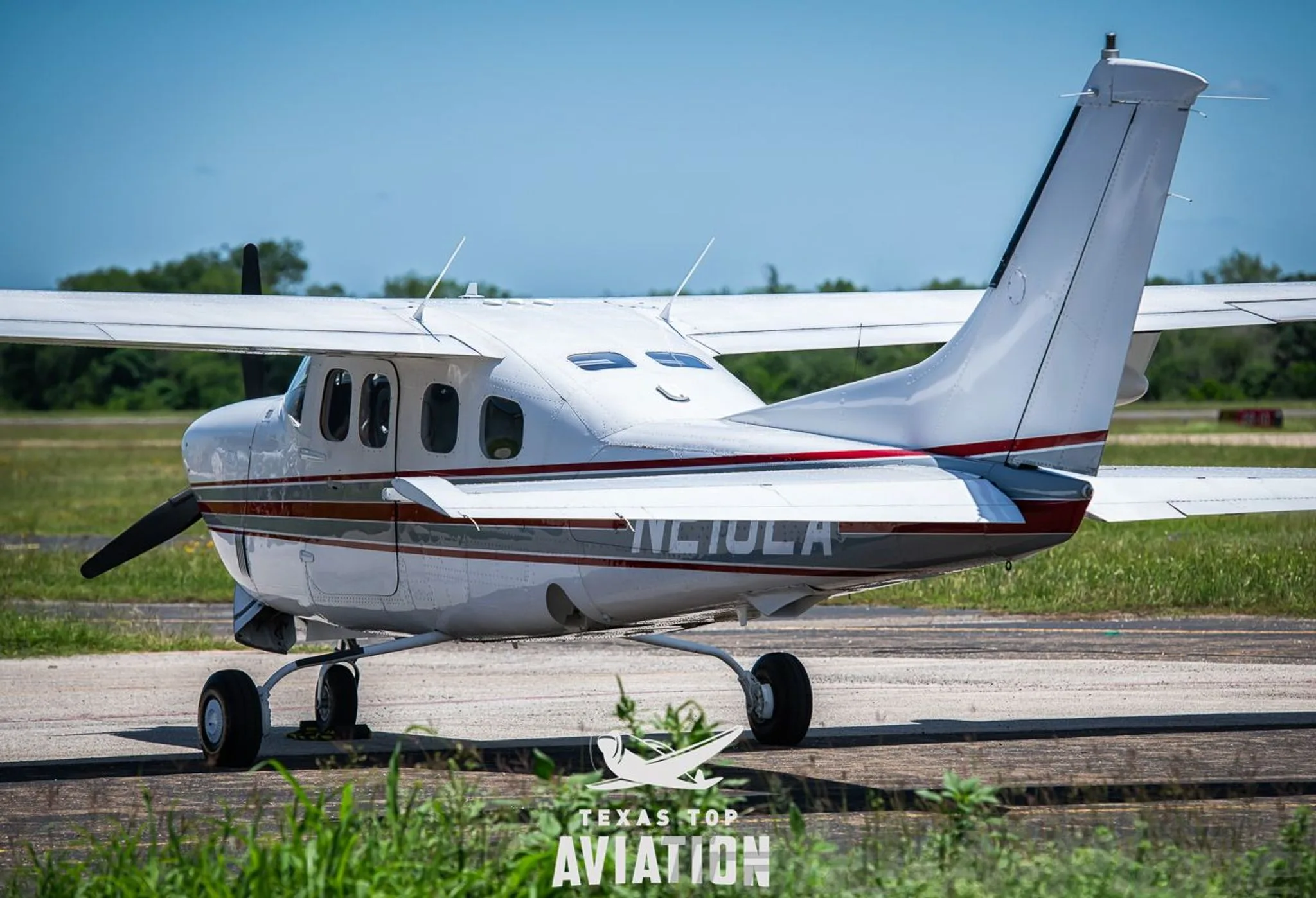 A white Cessna 210 Centurion (N210LA) with red and gray stripes, viewed from the rear-side on an airfield.