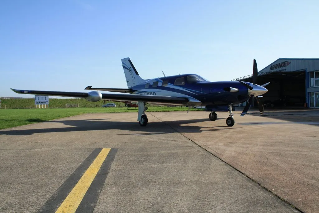 A front view of a blue and silver Piper M600SLS aircraft (G-VERO) parked on a tarmac with a hangar in the background.