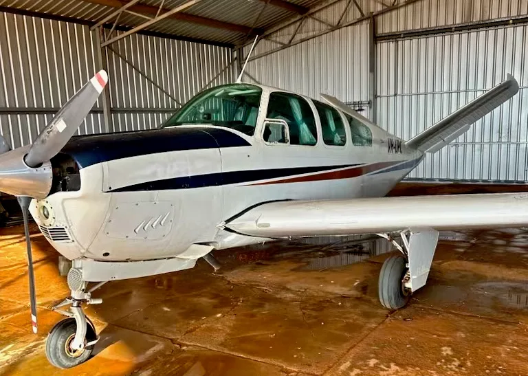 A white and blue Beechcraft Bonanza aircraft with a single propeller sits inside a hangar.
