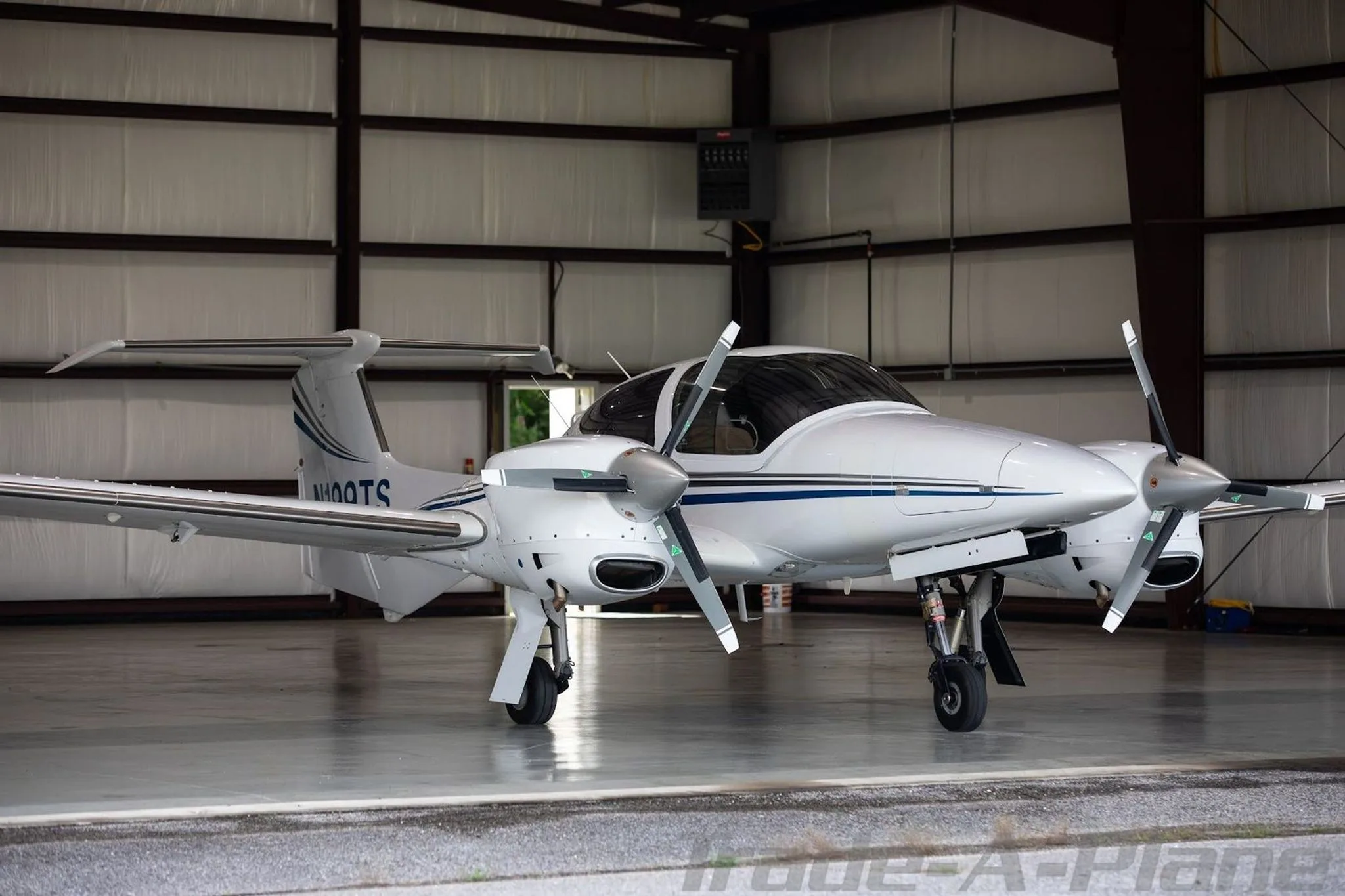 A white Diamond DA42-NG aircraft with blue and grey stripes is parked inside a hangar, viewed from the front-right.