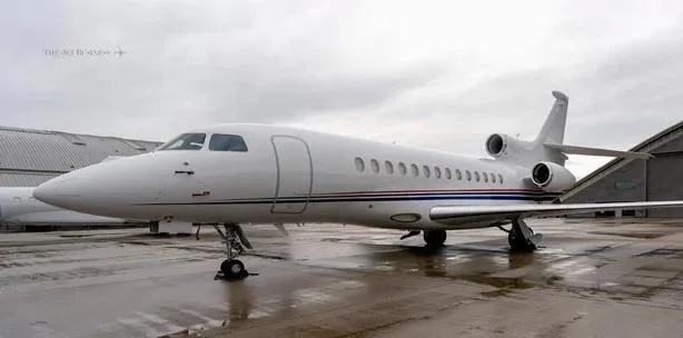 A white Dassault Falcon business jet with blue and red fuselage stripes, parked on a wet tarmac next to a hangar.