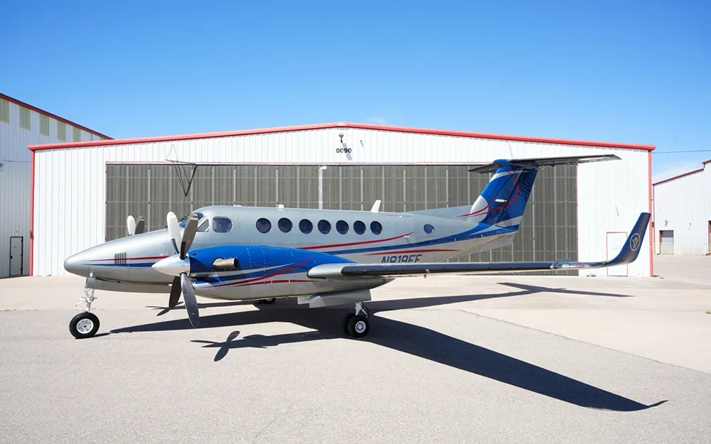 A silver and blue twin-engine turboprop aircraft (likely a King Air) parked on a tarmac in front of hangars under a clear blue sky.