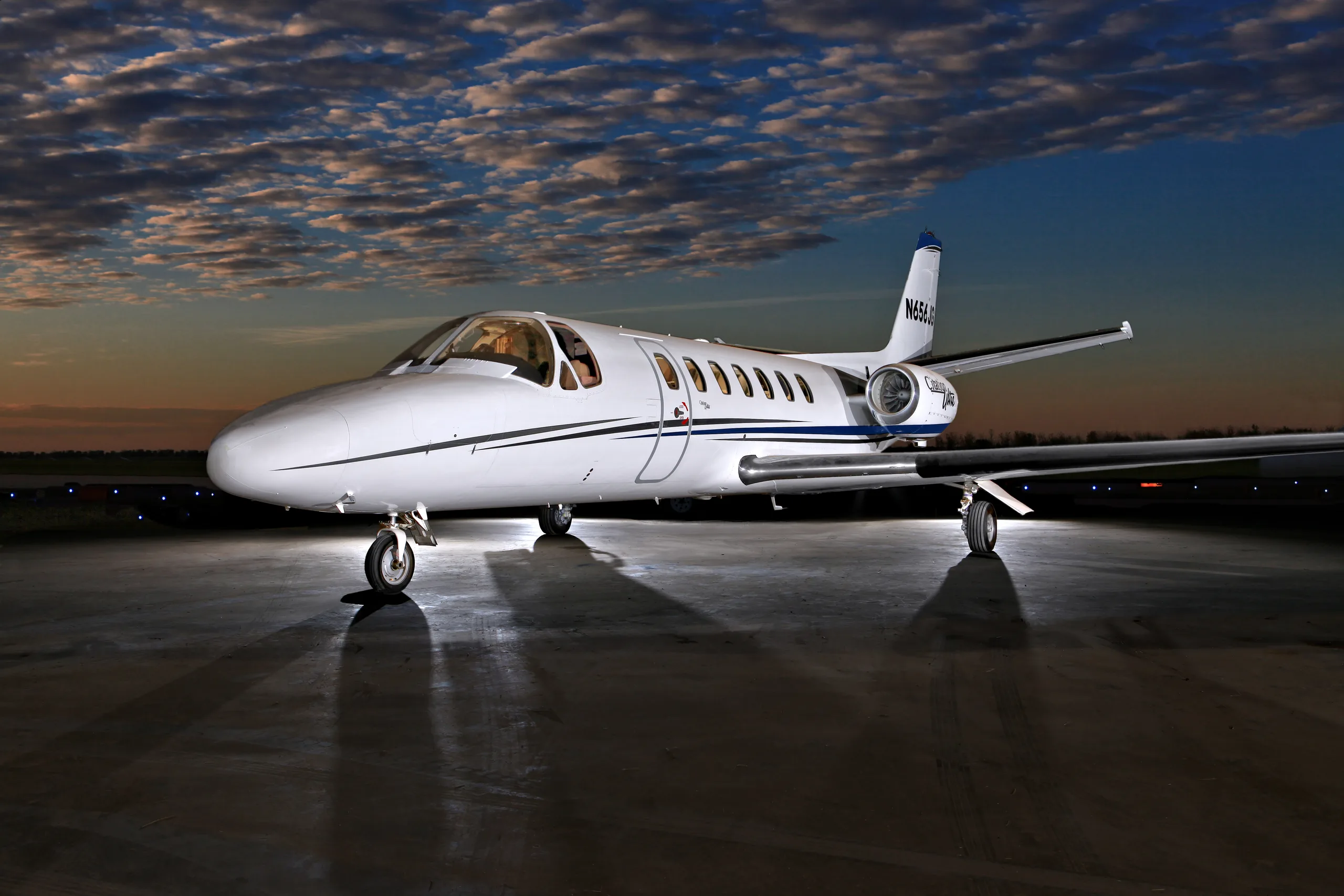 A white Cessna Citation Ultra business jet with blue stripes is parked on a tarmac at dusk, with runway lights in the background and a dramatic cloudy sky.