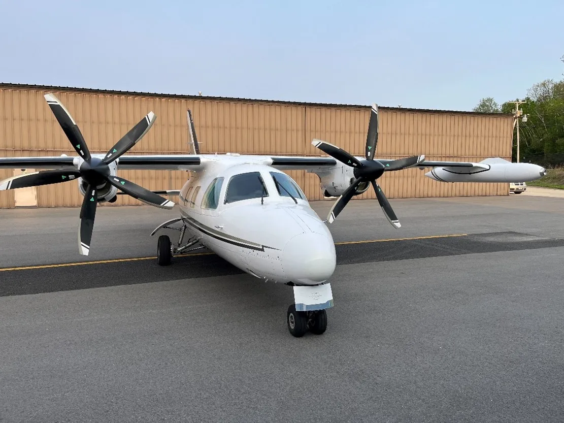 A white twin-turboprop aircraft with black propellers is parked on an asphalt apron in front of a light brown hangar.