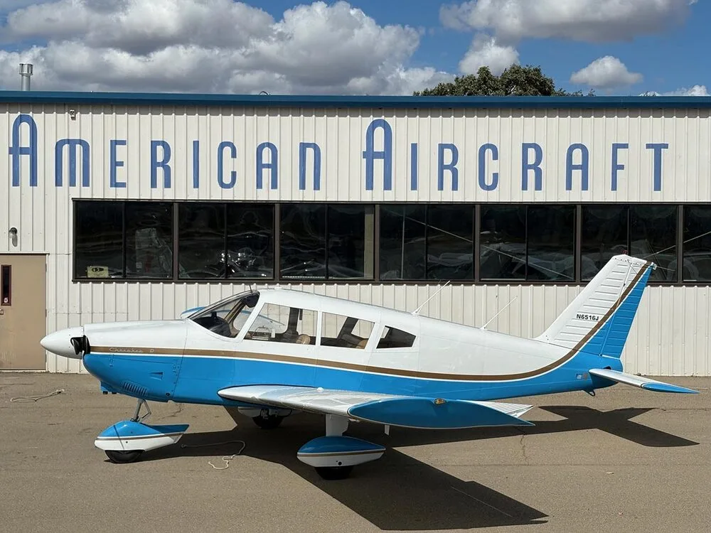 A side view of the white and blue Piper Cherokee D aircraft (N6516J) parked outside the "AMERICAN AIRCRAFT" building.