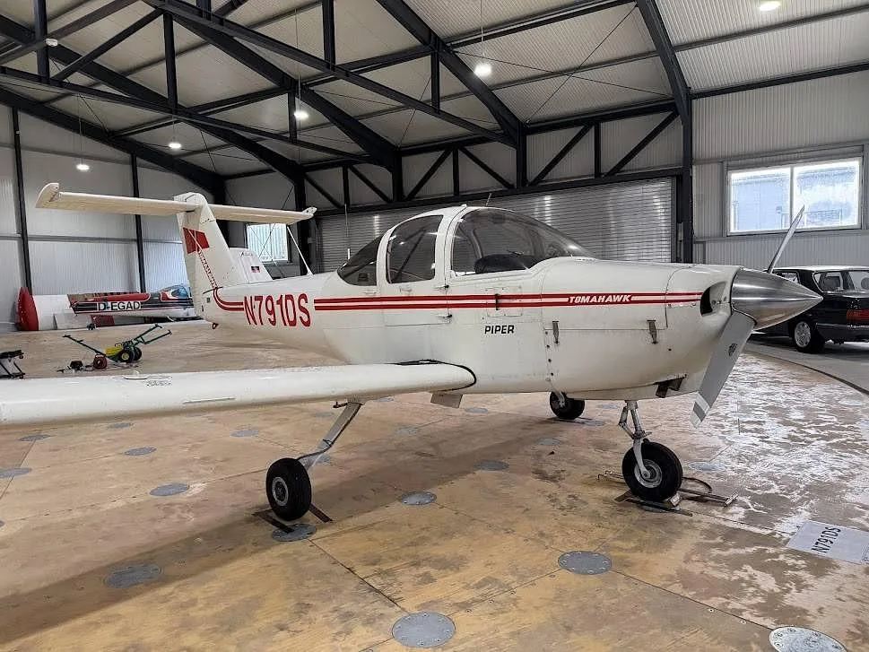 A white Piper Tomahawk aircraft with red stripes and registration N791DS is parked inside a hangar, viewed from the front-right.