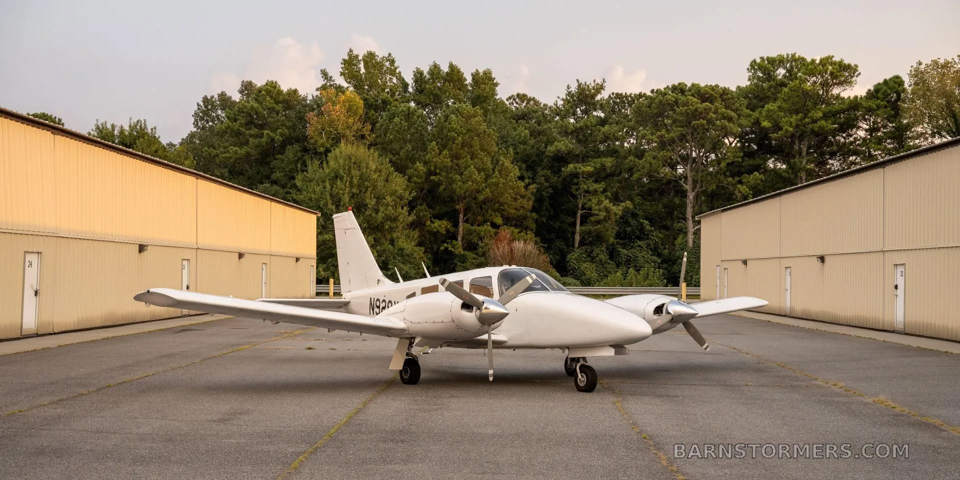 A white twin-engine propeller aircraft (N928X) is parked on an asphalt ramp between two hangars at sunset.