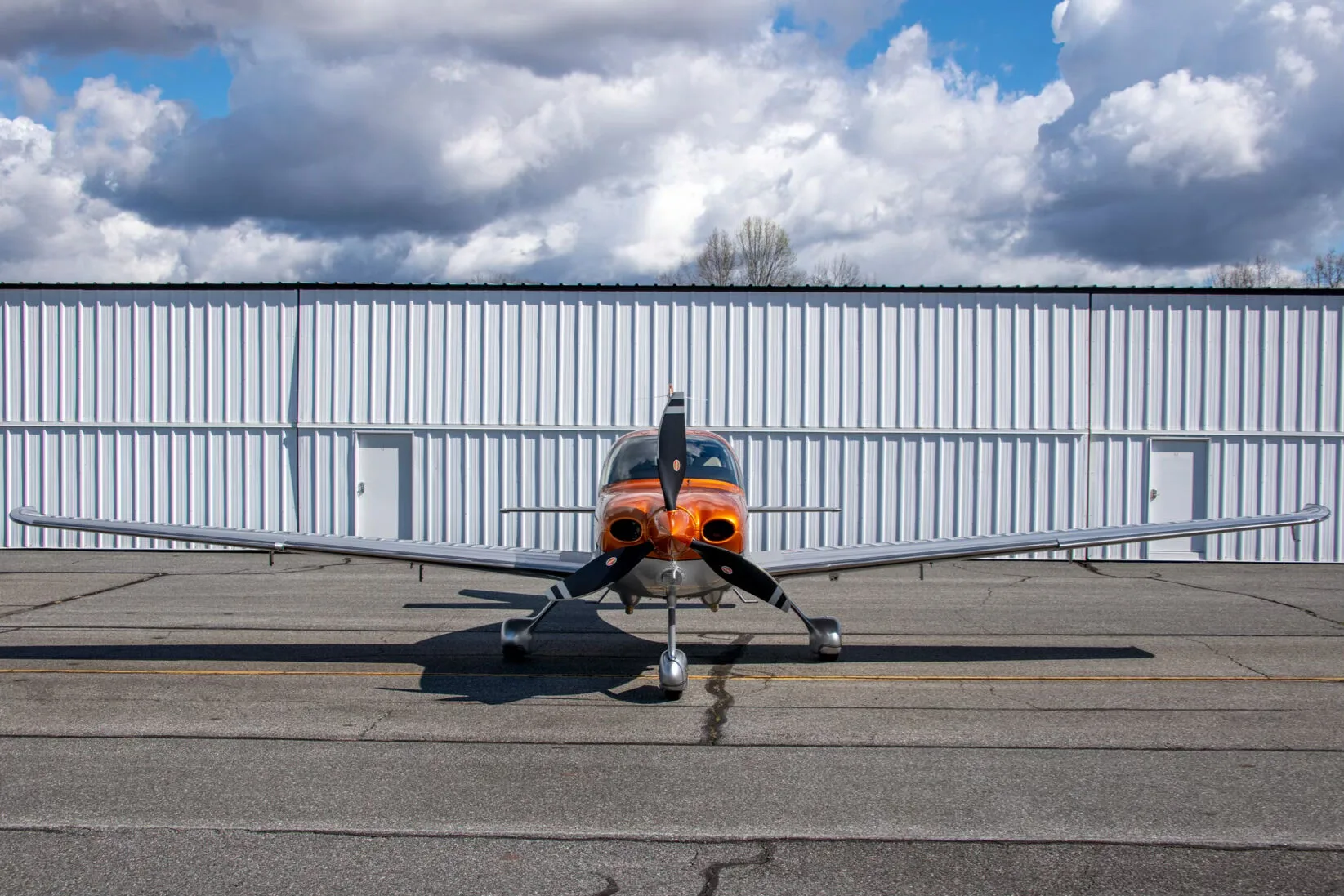 A head-on view of a metallic orange and silver Cirrus SR22 aircraft (N356HT) parked in front of a white hangar.