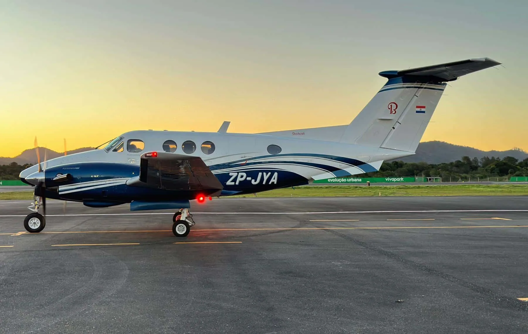 A blue and white Beechcraft King Air 350 turboprop aircraft with its navigation lights on is parked on an airfield at sunset.