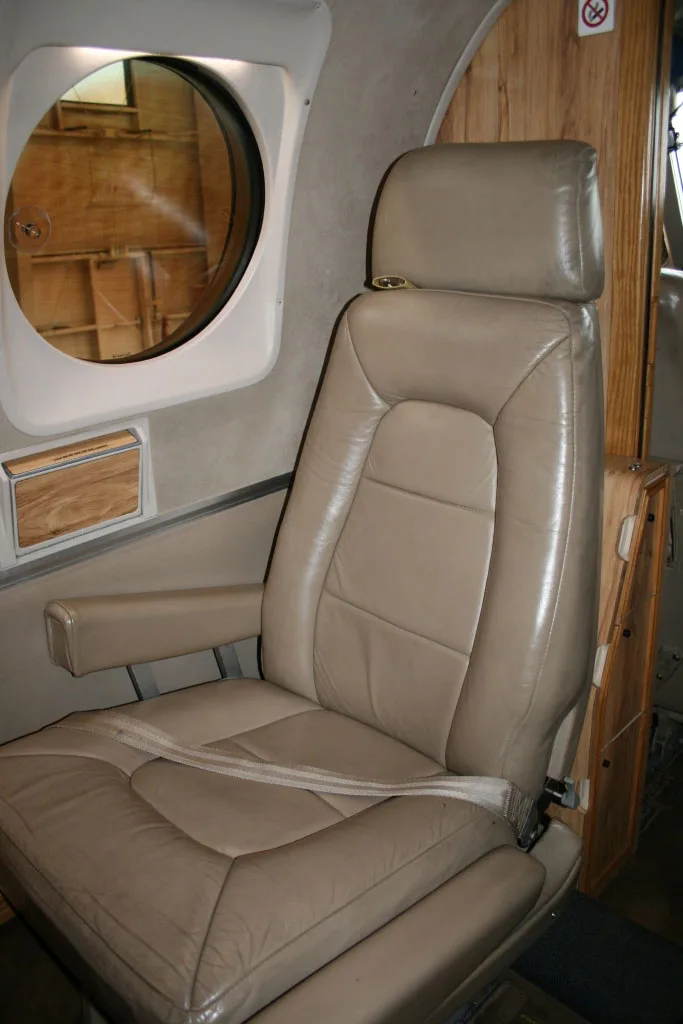 A close-up of a tan leather passenger seat in the aircraft cabin, next to a large oval window.