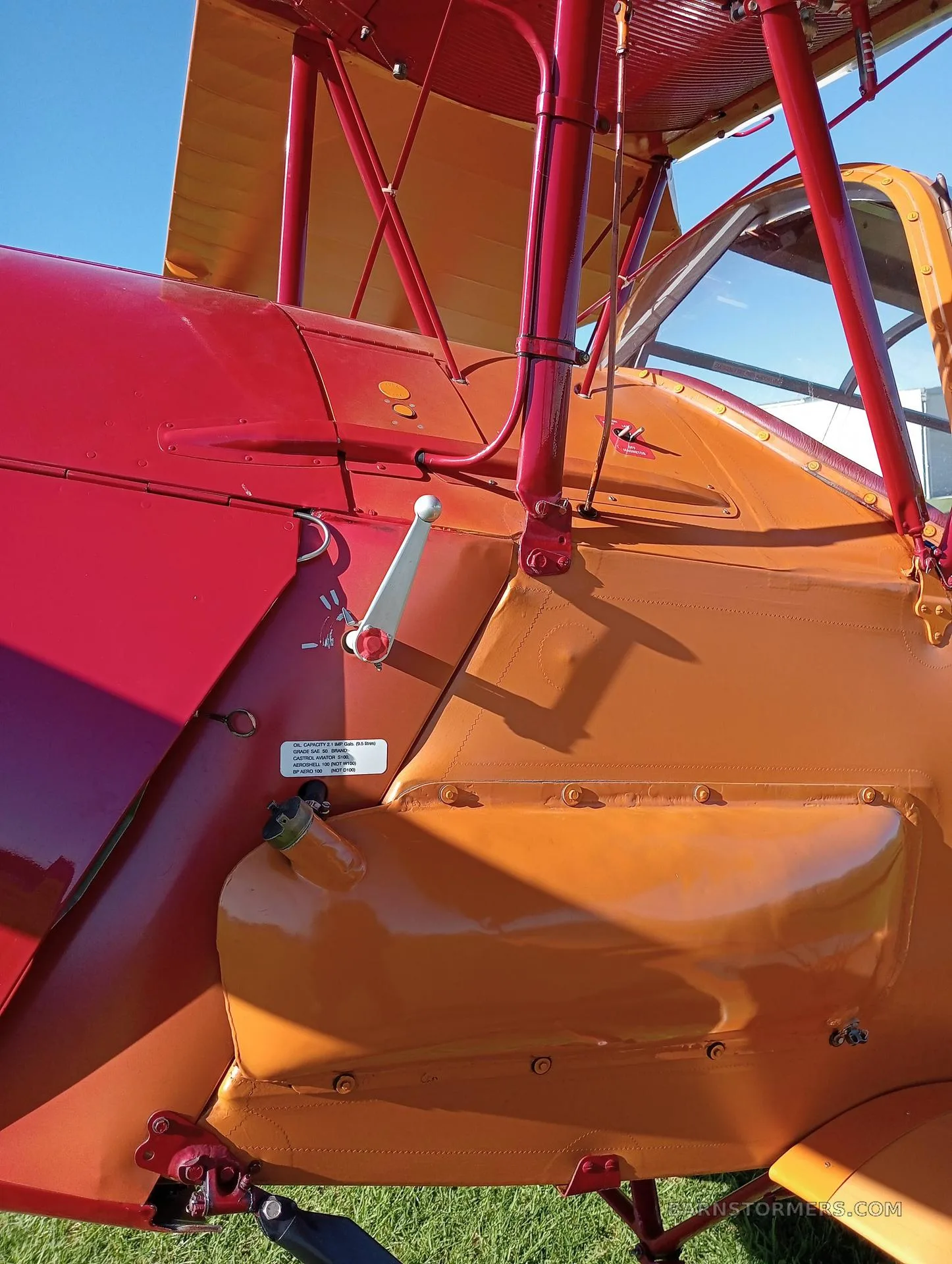 A close-up view of the red and orange fuselage and wing of a biplane, showing a fuel cap and a crank mechanism.