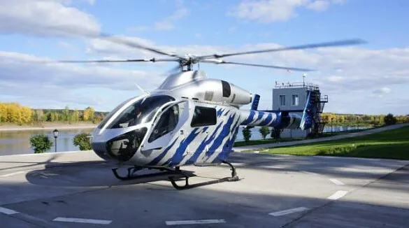 A silver and blue striped helicopter with its rotors spinning, parked on a helipad next to a river.