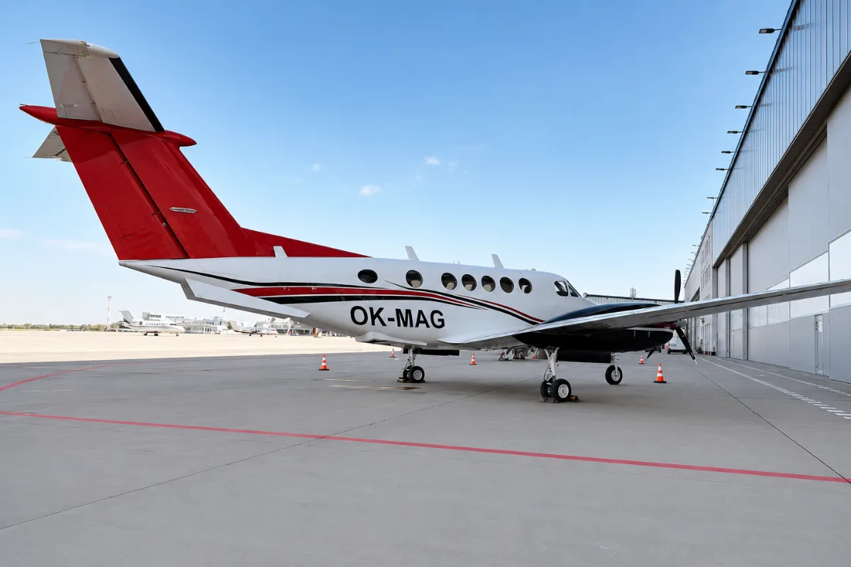 A white and red twin-engine turboprop aircraft with registration OK-MAG parked on the tarmac next to a hangar.