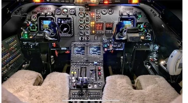 The illuminated cockpit of a business jet, featuring multiple screens, analog gauges, and control yokes with sheepskin covers.