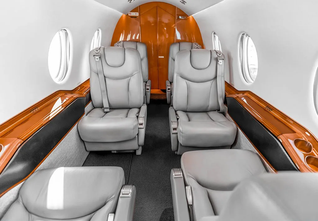 The aircraft cabin features gray leather seats and wood trim, looking towards the rear of the cabin.