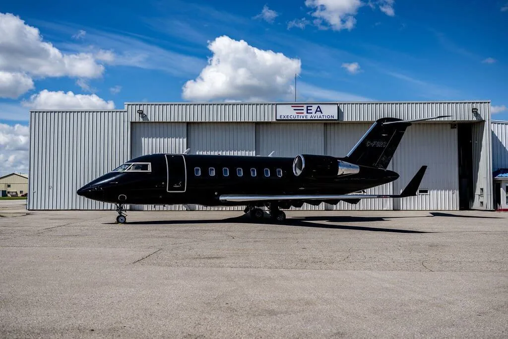 A black Bombardier Challenger 604 business jet with registration G-FWIG parked on an tarmac in front of a hangar under a blue sky.