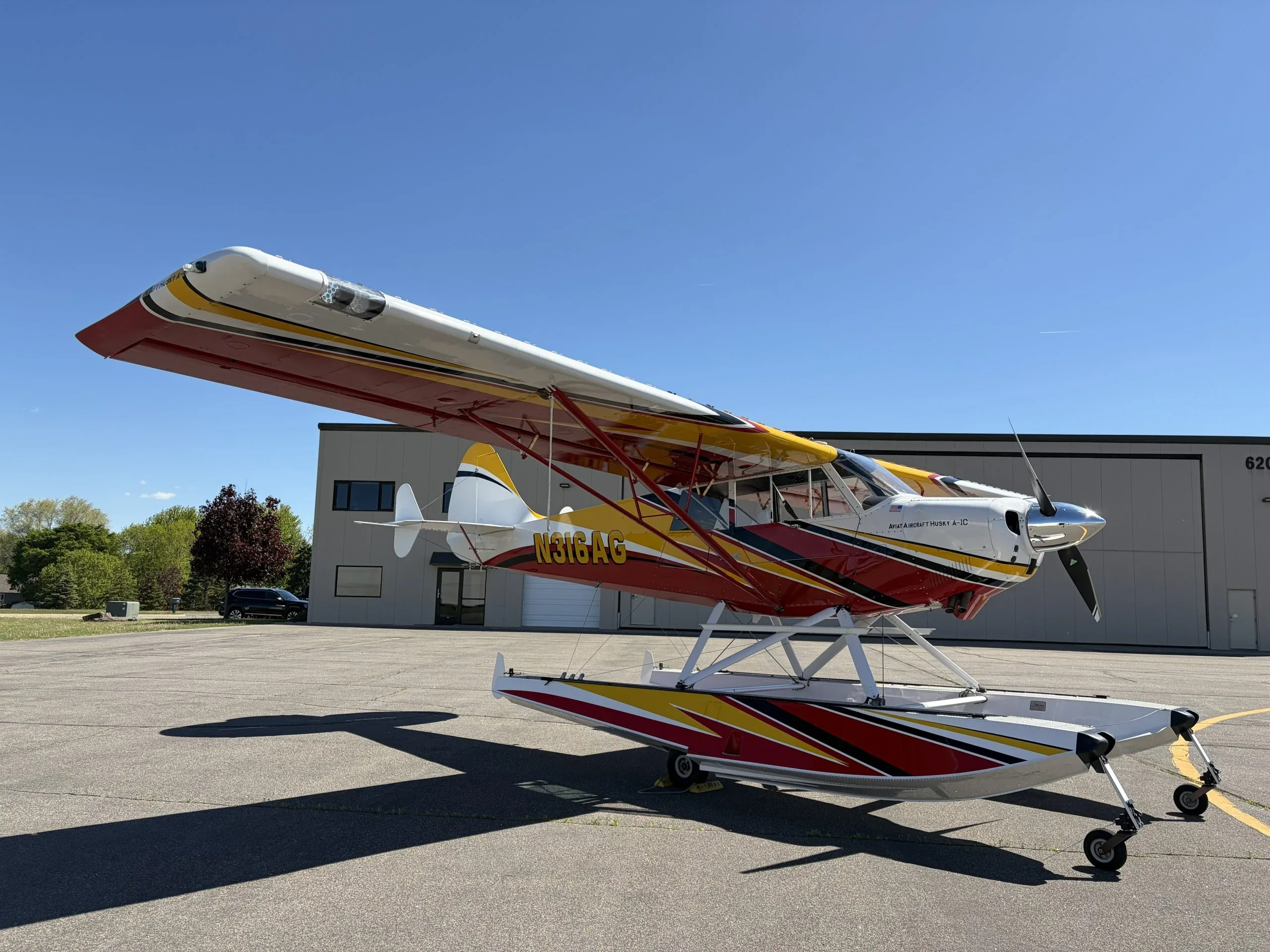 A three-quarter front view of the red, yellow, and white Aviat Aircraft Husky A-1C seaplane, N316AG, on an airfield.