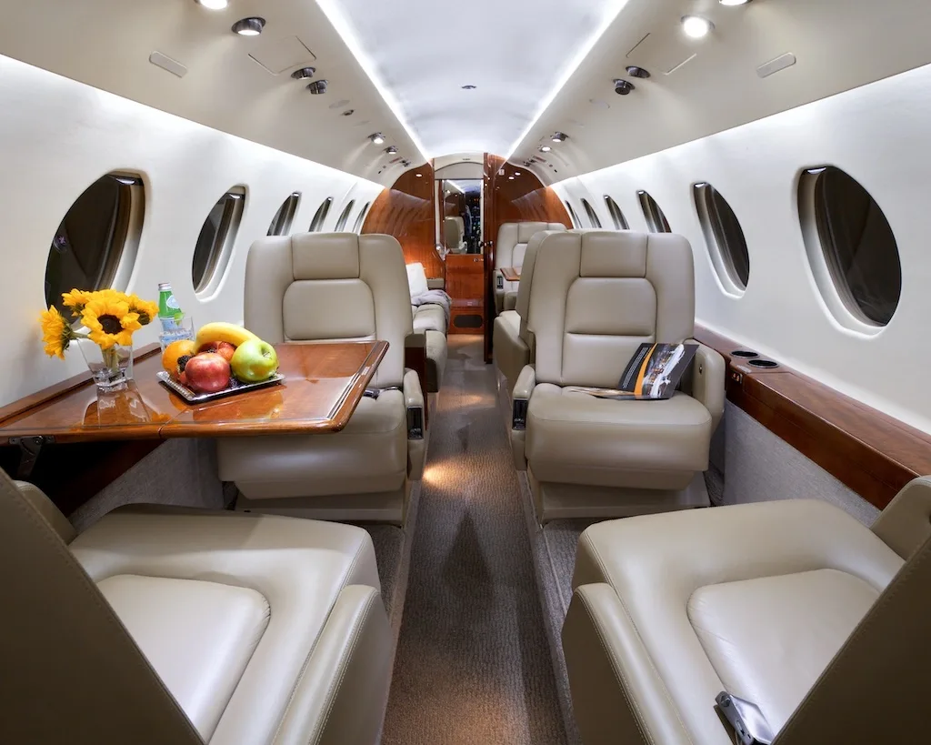 The luxurious cabin interior of a private jet, featuring beige leather seats, a wooden table with fruit and flowers, and large windows.