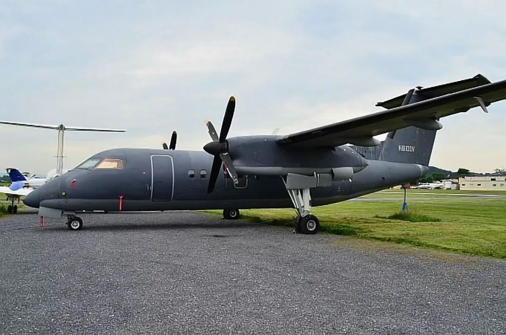 A dark gray turboprop aircraft, N8100V, is parked on an airfield tarmac with grass in the background.