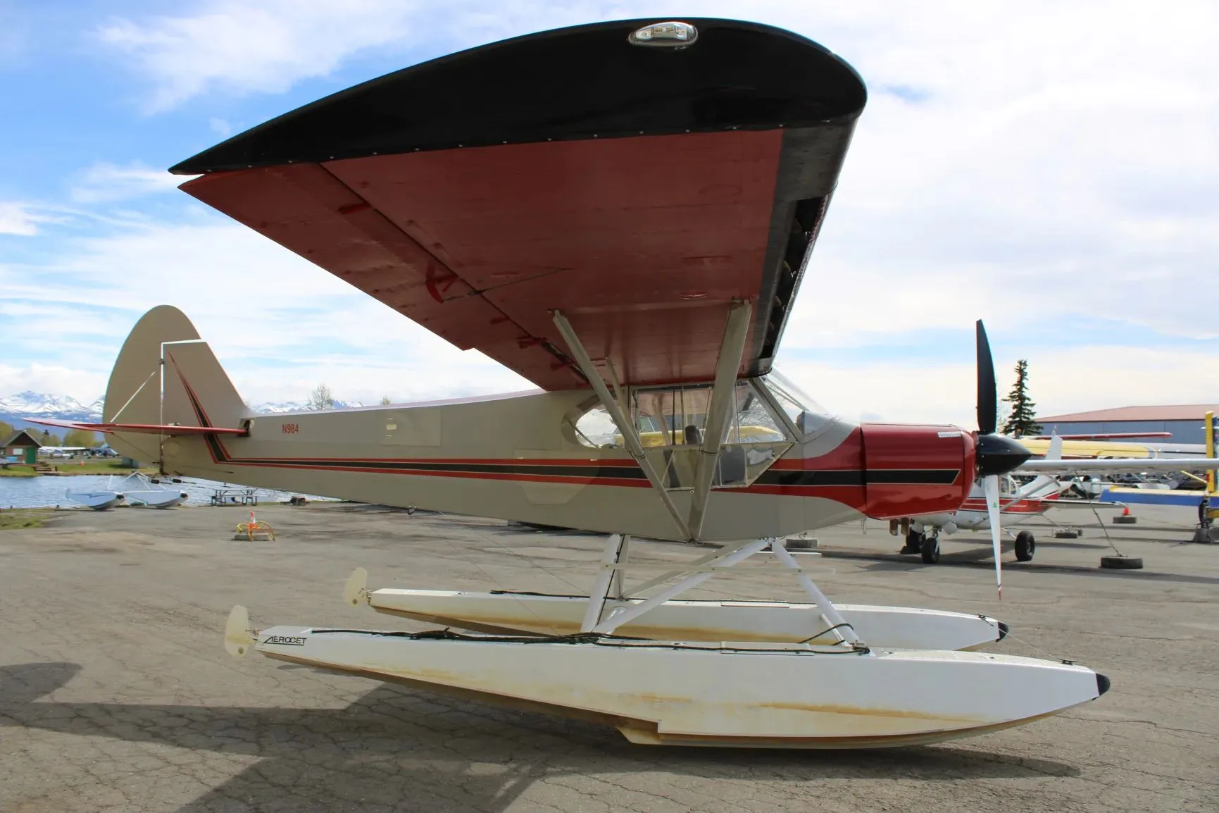 A side view of a red and grey floatplane with N984 on the tail, parked on an apron with other aircraft in the background.