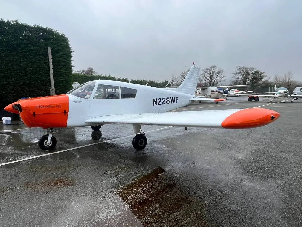 A white and orange Piper Cherokee 140 (N228WF) parked on a wet tarmac at an airfield, viewed from the front-left.