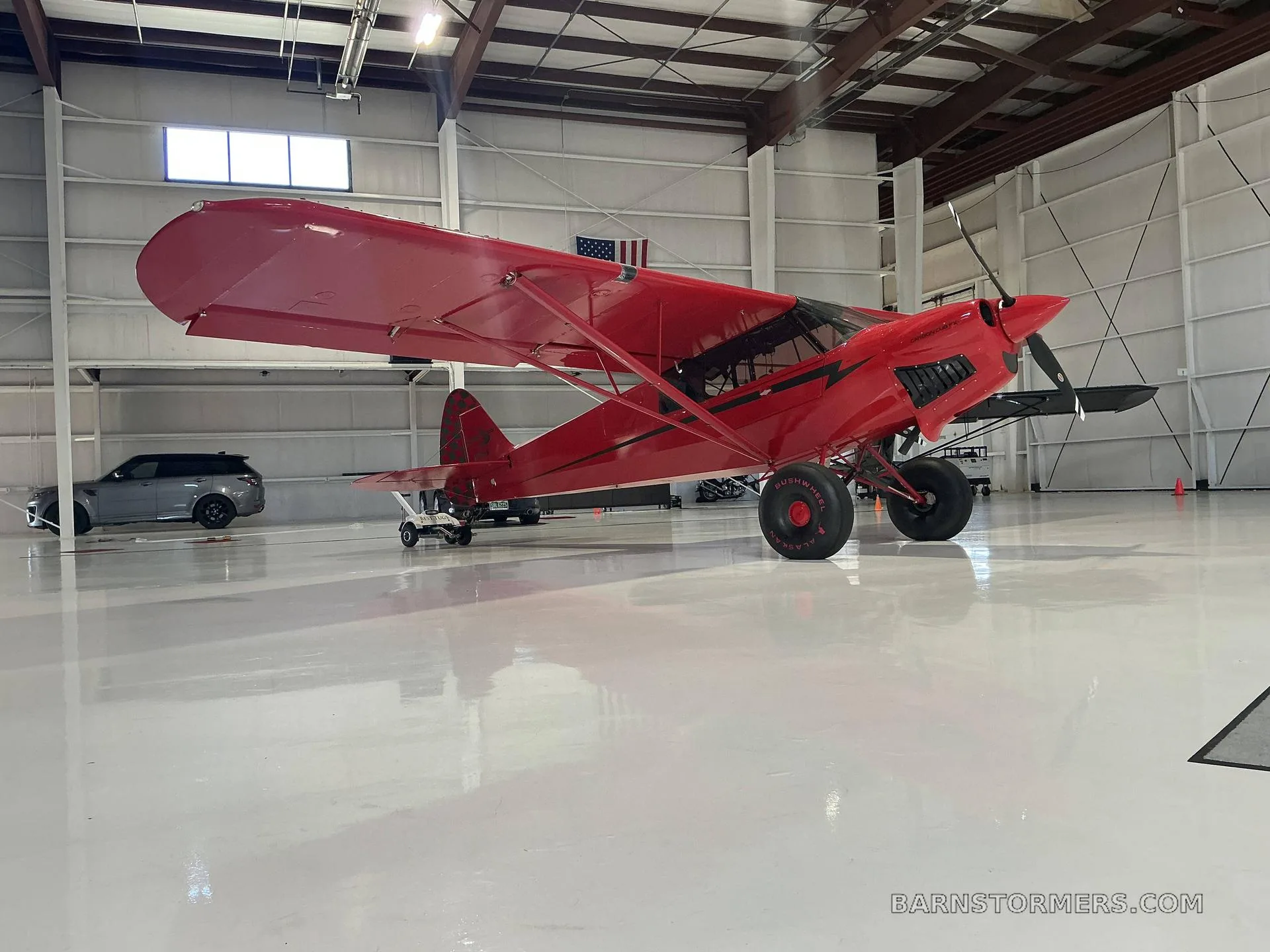 A red Bushmaster aircraft with large tundra tires is parked in a hangar, viewed from the front-right.