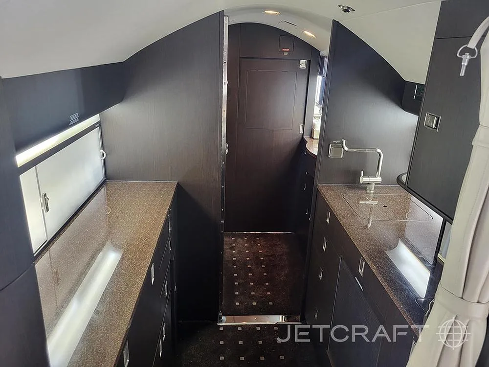 The galley area of a private jet, with dark wood cabinetry, a sink, and a light-colored countertop.
