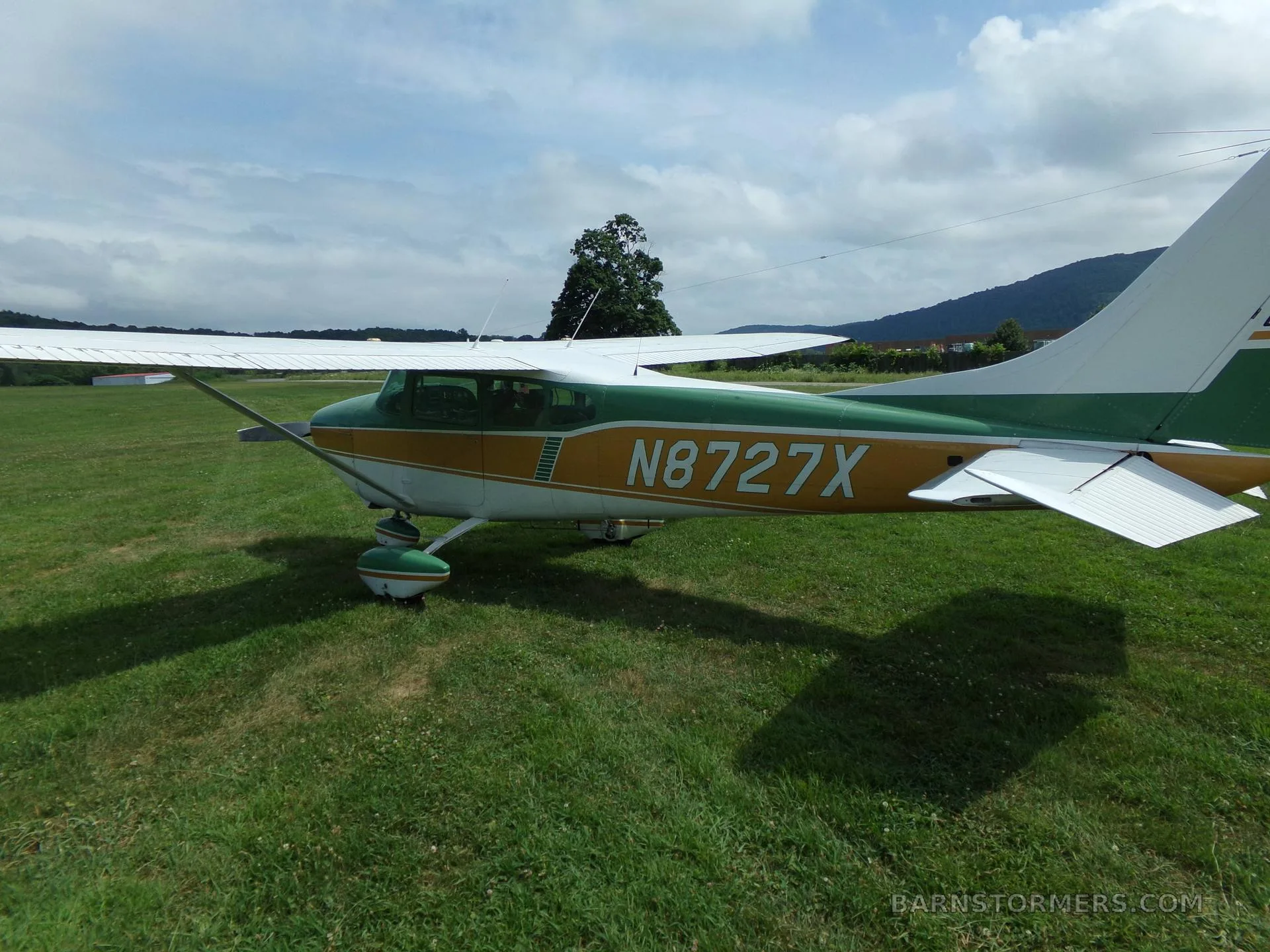 A green, gold, and white Cessna 172 aircraft with registration N8727X, viewed from the rear-left on a grassy field under a partly cloudy sky.