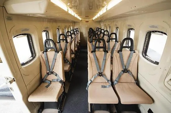 The interior cabin of an aircraft, showing rows of beige passenger seats with shoulder harnesses.