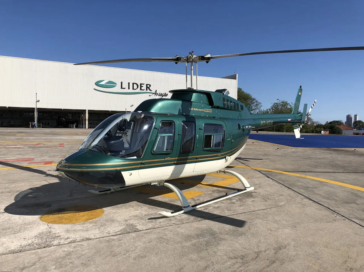 A green and white Bell helicopter (PT-YPY) is parked on a tarmac in front of a LIDER Aviação hangar under a clear blue sky.