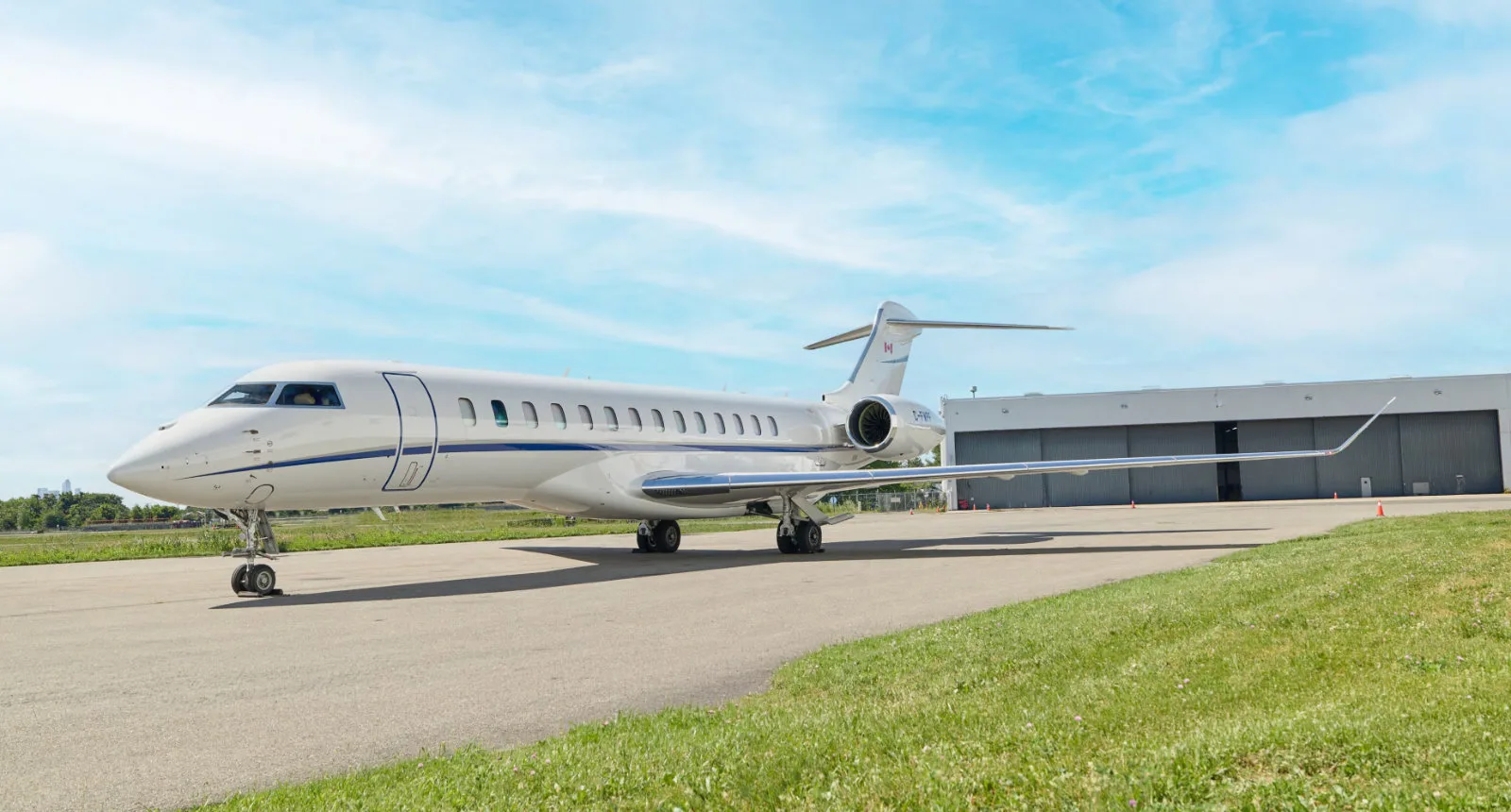 A white Bombardier Global business jet with blue stripes is parked on an airport tarmac with a hangar in the background.