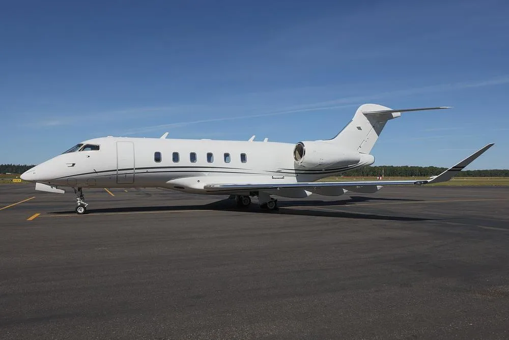 A white Bombardier Challenger business jet with black stripes on the tail and winglets, parked on an asphalt tarmac under a clear blue sky.