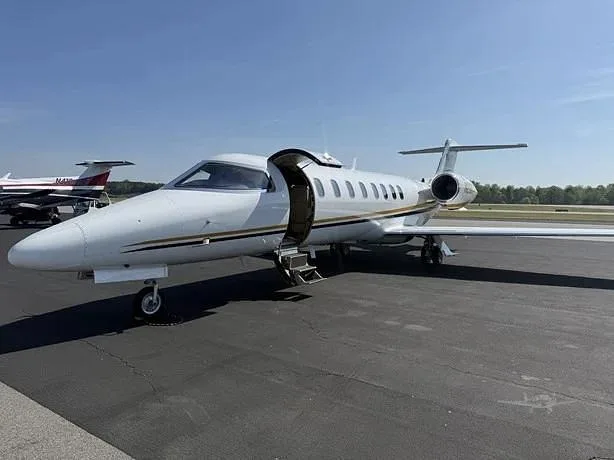 A white business jet with gold and black stripes, registration N26CT, parked on an asphalt tarmac with its main door open and stairs extended.