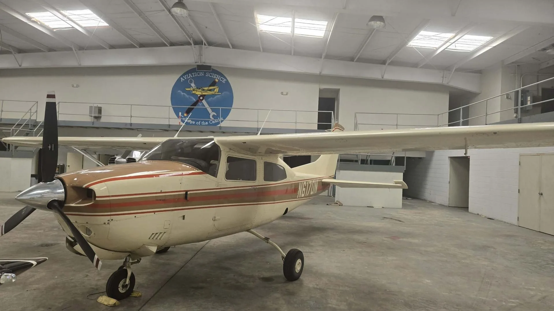 A front-side view of the N6170N aircraft, a white and brown high-wing single-engine plane, parked in a hangar with an "Aviation Science" mural.