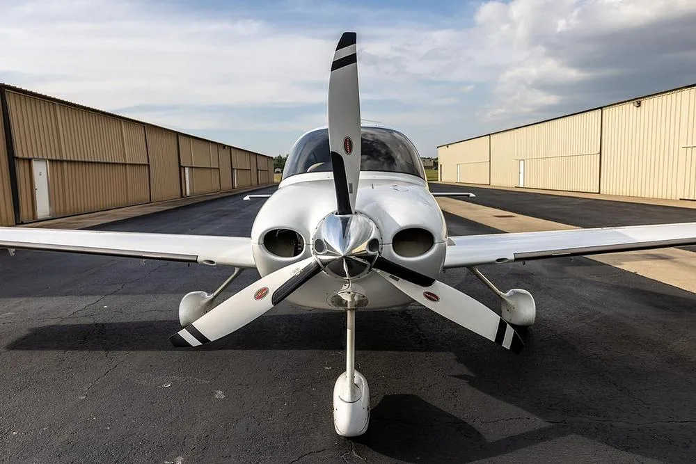 A head-on view of a white Cirrus SR22T aircraft with a three-bladed propeller, parked on an airfield between hangars.