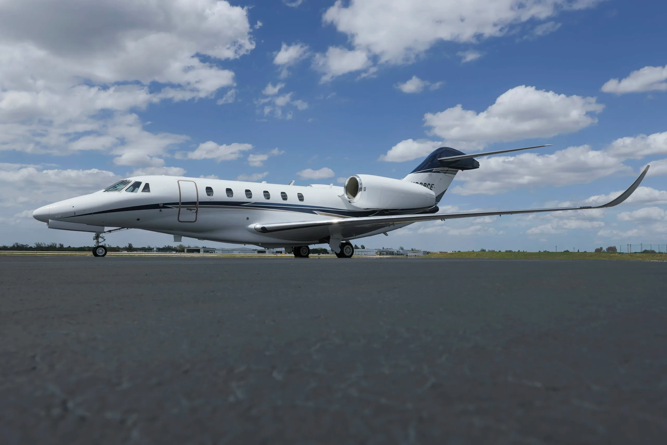 A white business jet with blue and grey stripes on the tail and fuselage is parked on an asphalt tarmac under a partly cloudy sky.