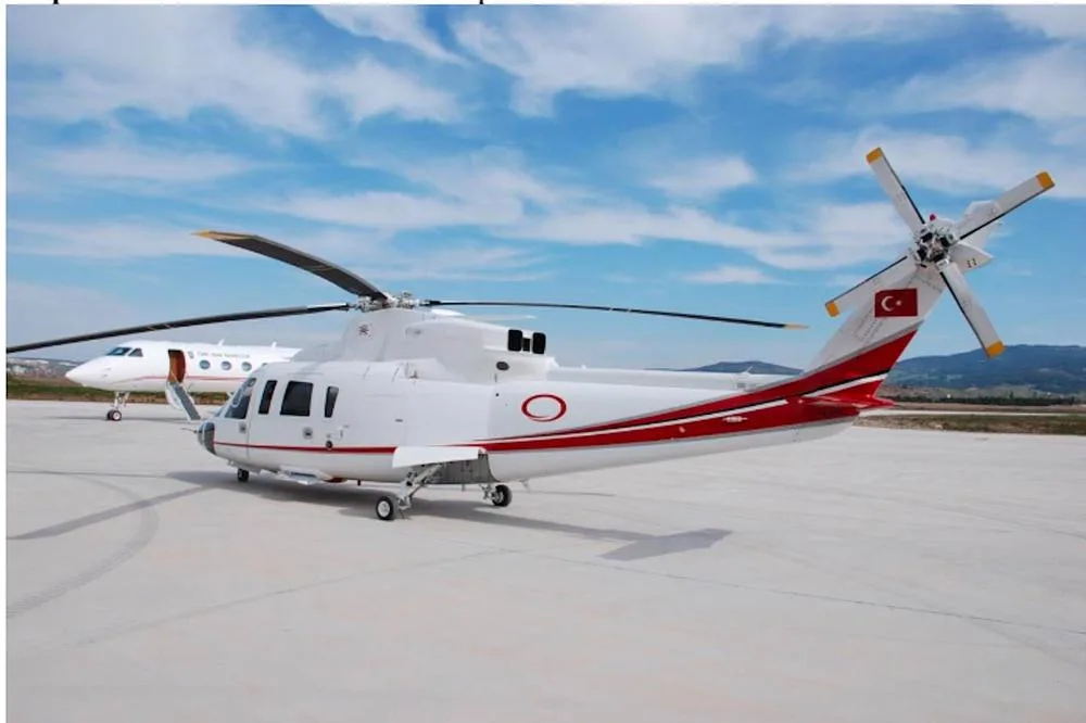 A white helicopter with red stripes and a Turkish flag on the tail, parked on an airfield next to a white business jet under a blue sky.