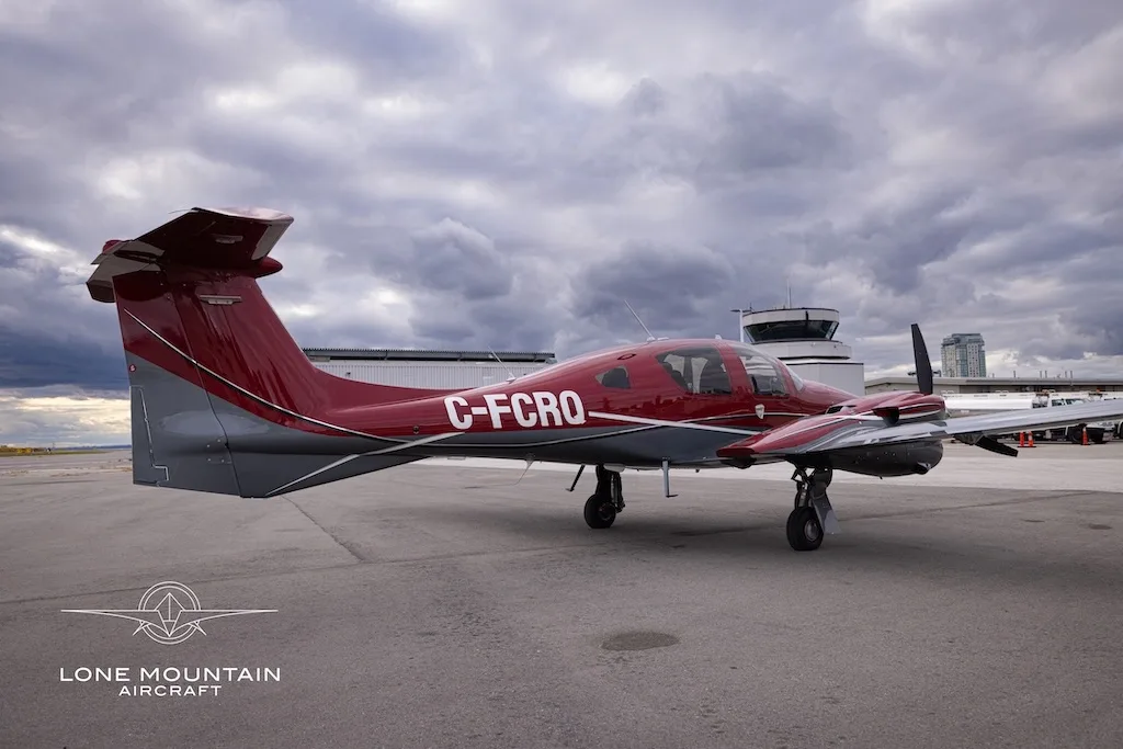 A red and grey Diamond DA62 aircraft with registration C-FCRQ parked on an airfield with a control tower in the background.