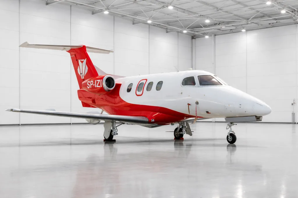A white and red Embraer Phenom 300, registration SP-IZI, parked inside a modern hangar.