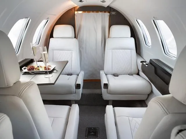 The elegant white leather interior of a private jet cabin, featuring a table with champagne and snacks between two seats.