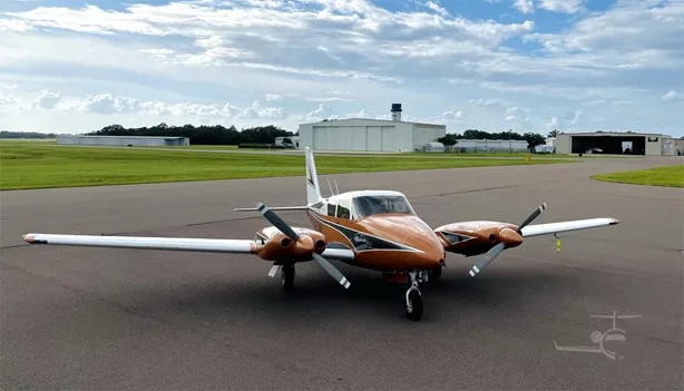 Front view of a copper and white Piper Comanche twin-engine aircraft on an airfield.