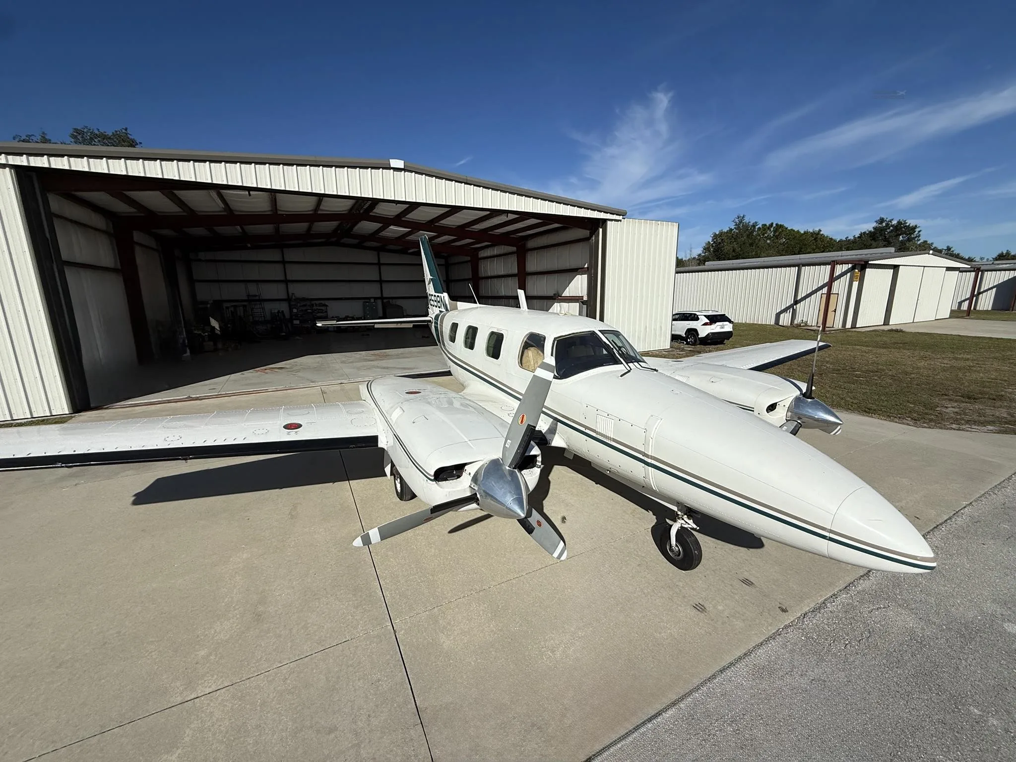 A white twin-engine propeller aircraft with a green stripe is parked on a tarmac in front of an open hangar under a blue sky.