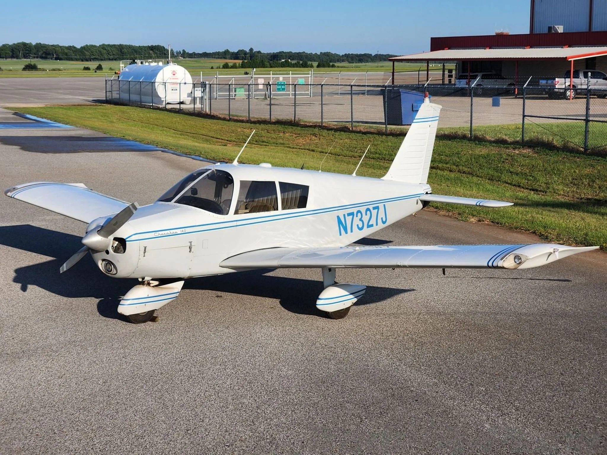 A white Piper Cherokee 140 aircraft, N7327J, with blue stripes, parked on an airport tarmac on a sunny day.