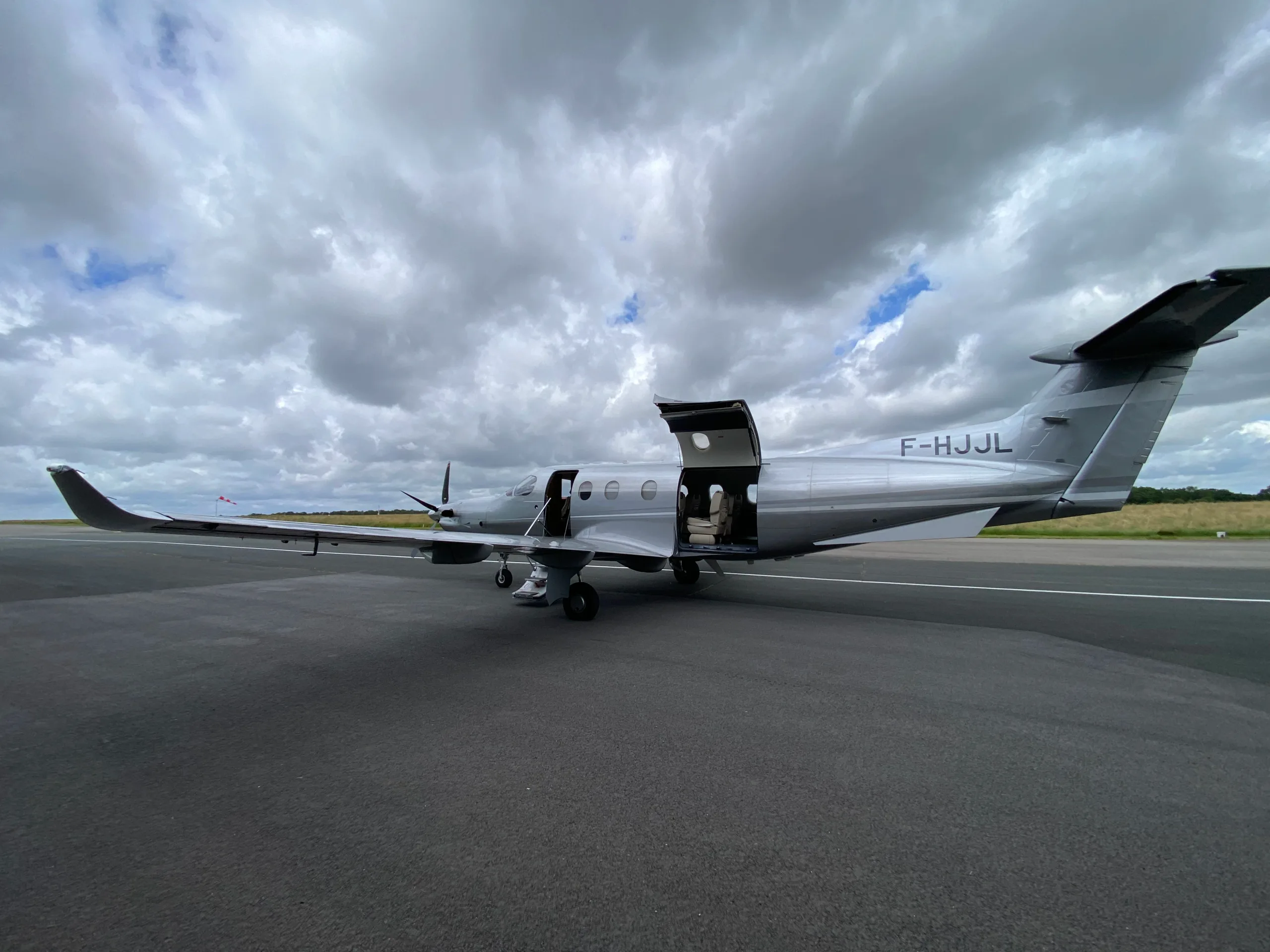 A silver Pilatus PC-12 aircraft with its main and cargo doors open, viewed from the rear-left on an asphalt runway under a cloudy sky.