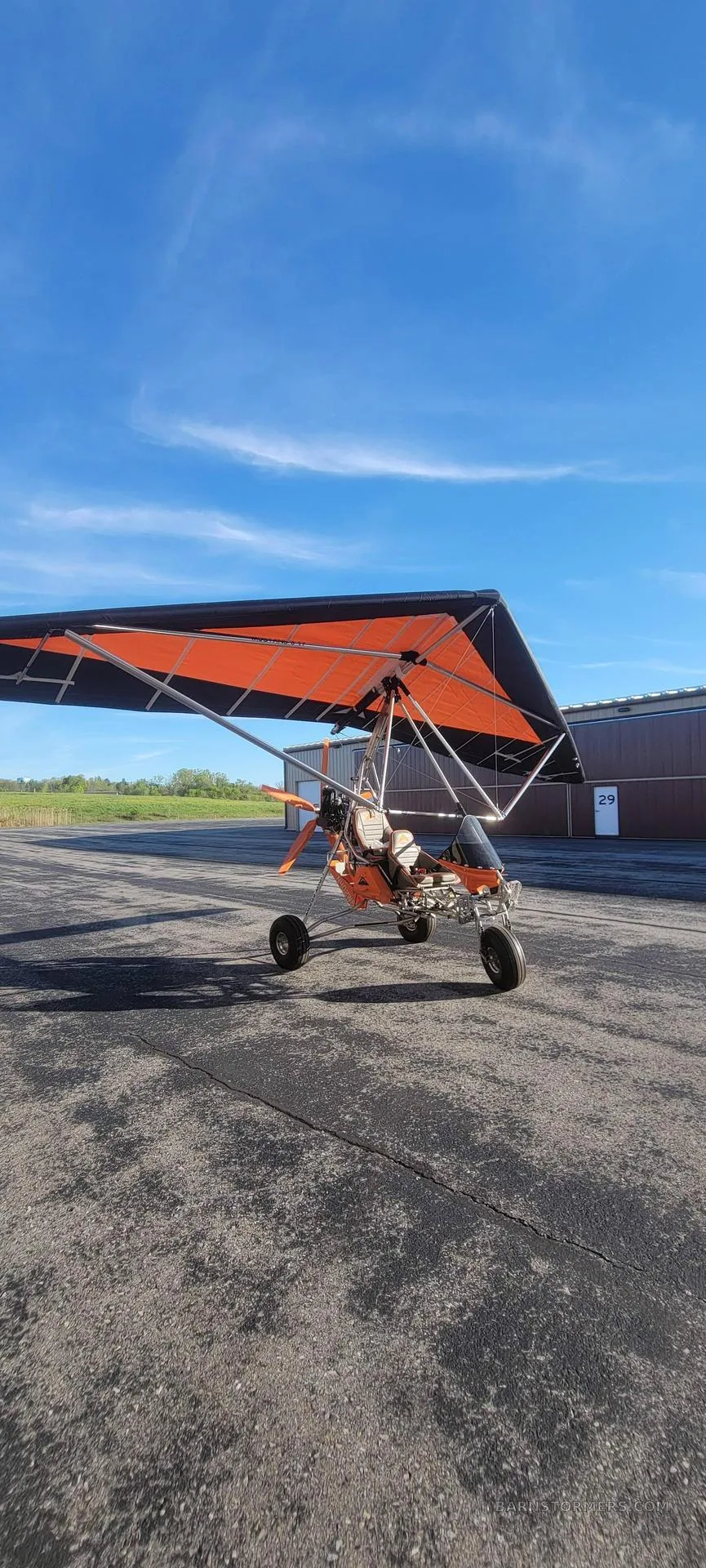 An orange and black weight-shift control ultralight aircraft parked on an airfield tarmac under a blue sky.