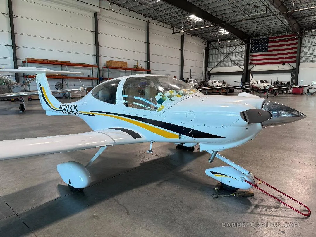 A white Diamond DA40 aircraft with yellow and black stripes, parked in a hangar with an American flag in the background.