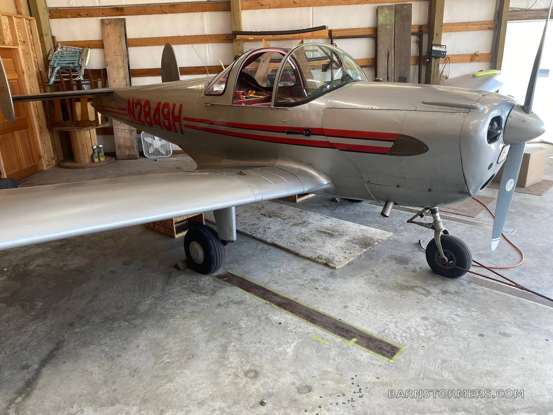 A silver Ercoupe aircraft with red stripes and tail number N2849H parked inside a wooden-framed hangar.