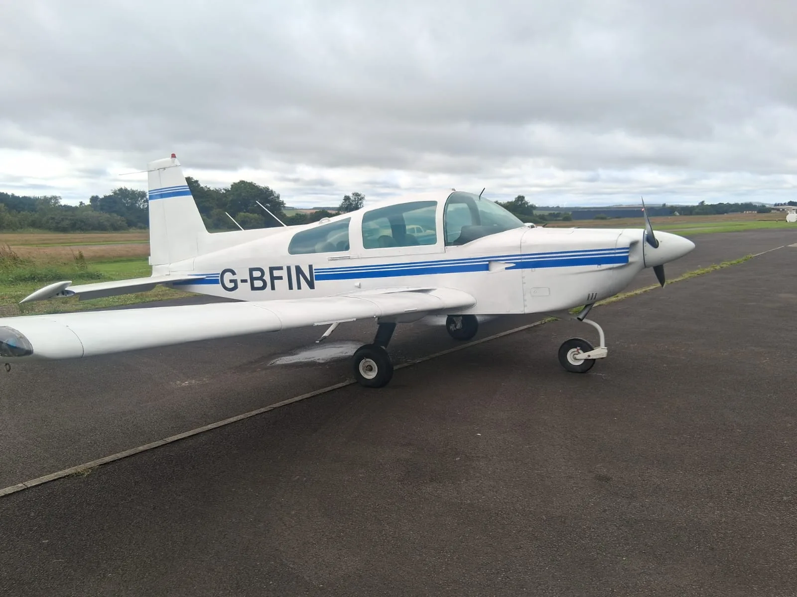 A white single-engine propeller aircraft with blue stripes, registration G-BFIN, parked on an airfield tarmac under a cloudy sky.